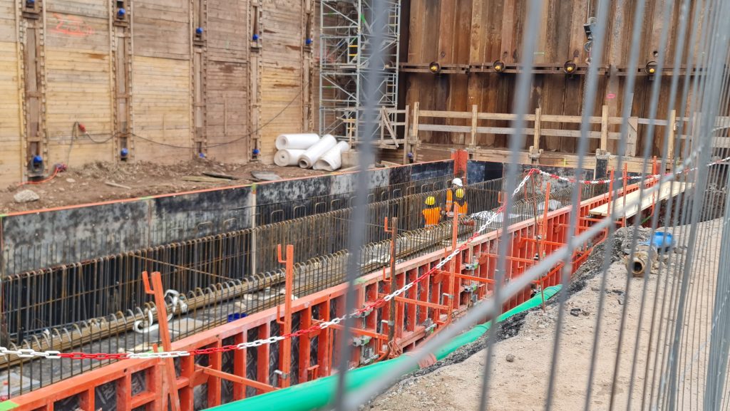 Bauarbeiter in tiefem Graben, Umzäunung im Vordergrund.